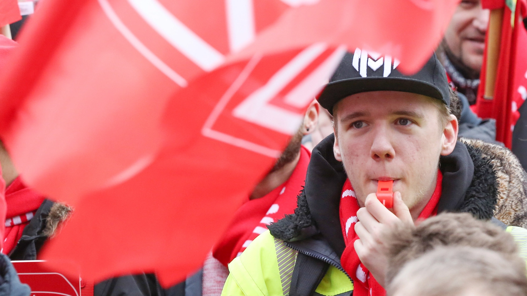 Junger Mann zwischen IG Metall-Fahnen pustet bei einem Warnstreik während der Tarifrunde in eine Trillerpfeife.