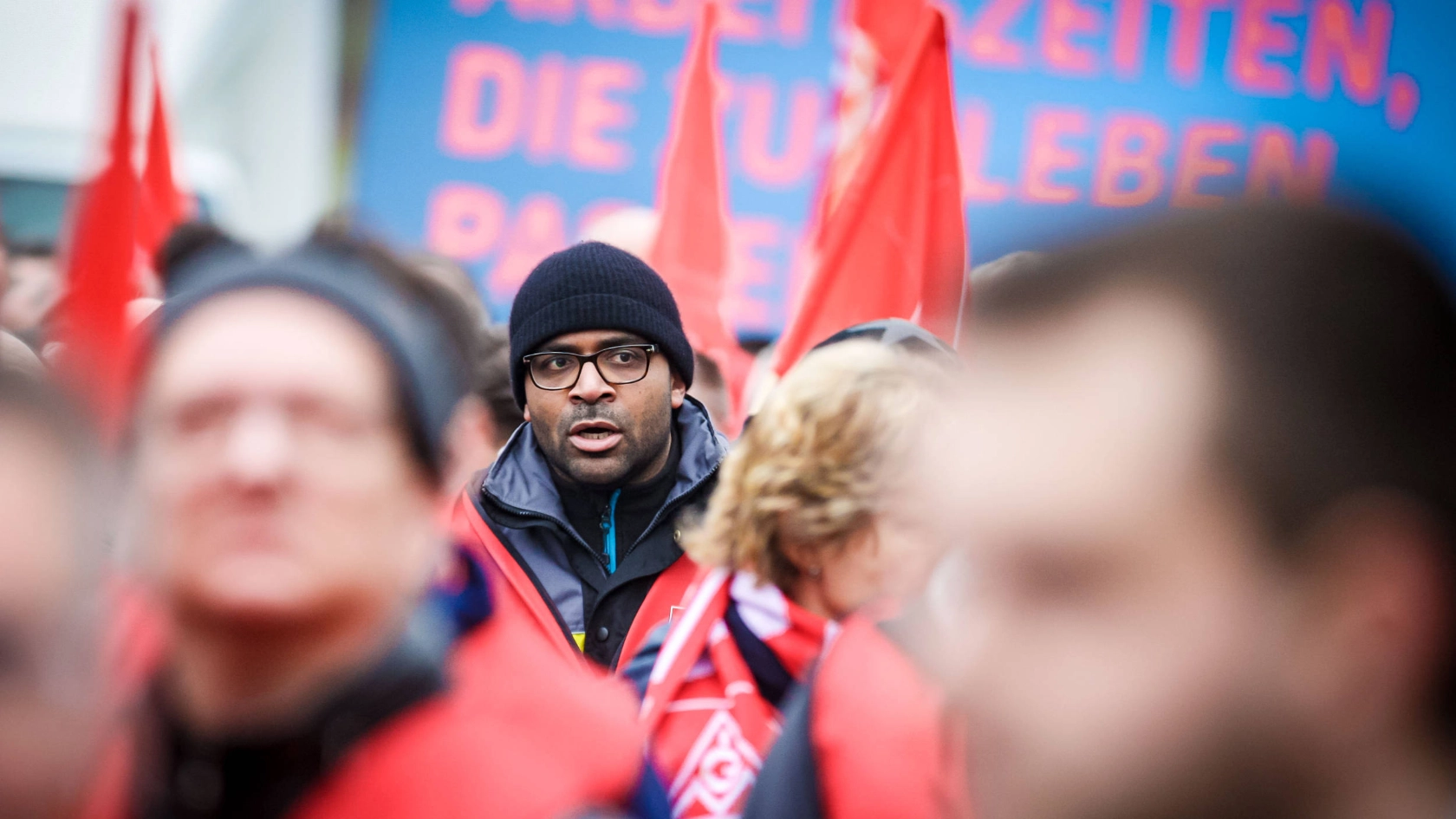 Ein Mann mit schwarzer Brille und Mütze steht in einer Menschenmenge.