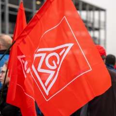 IG Metall-Fahne - im Hintergrund Beschäftigte bei einem Warnstreik 