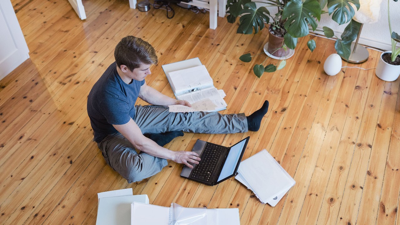 Junger Mann sitzt mit Notebook auf Fußboden und sortiert Papier und Akten
